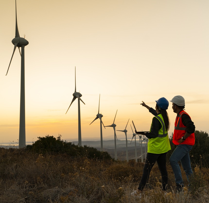 Man and woman in high vis outside pointing at windfarm against orange sky